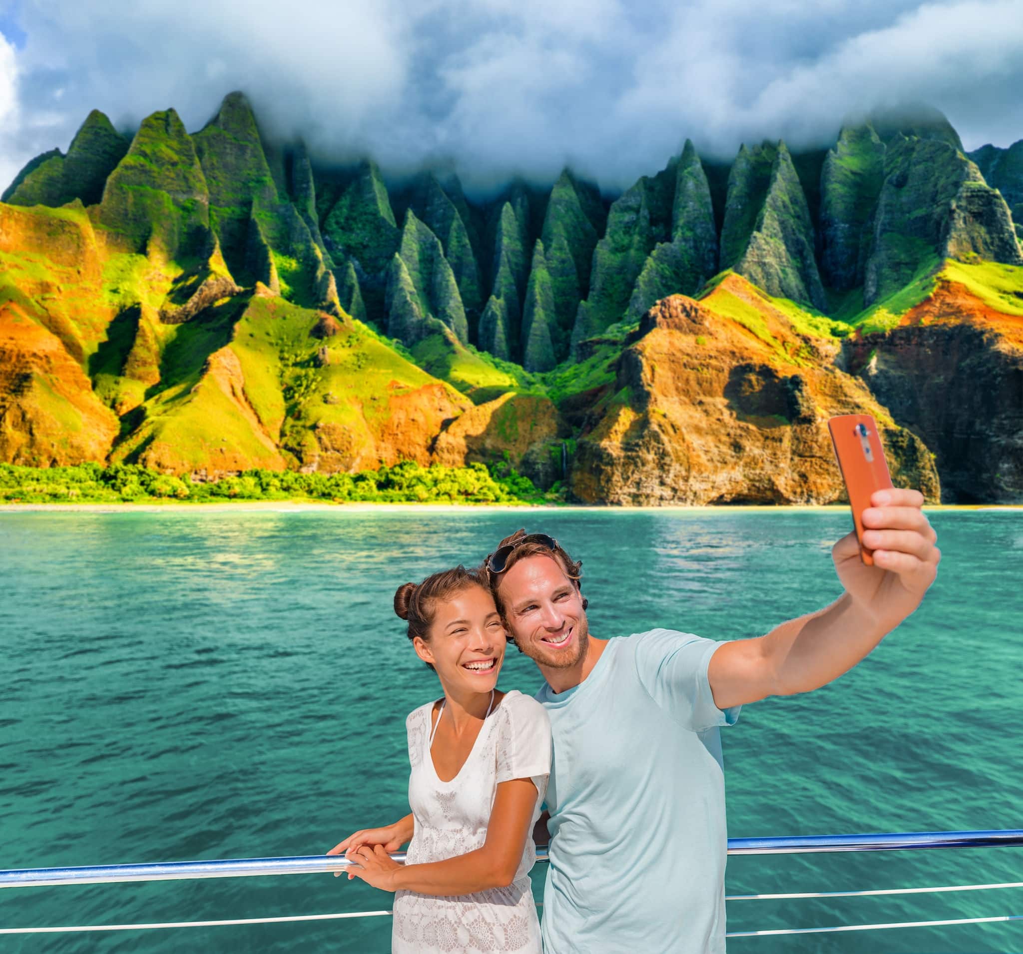 Hawaii cruise ship couple tourists taking selfie photo with phone at Na Pali Coast, Kauai island, USA summer vacation travel. Happy Asian woman, Caucasian man, interracial friends group of people.