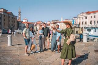 Mixed-race female tour guide leads a diverse group of travelers through a small Mediterranean town. They are actively exploring the streets, immersing themselves in the local culture and enjoying the experience.