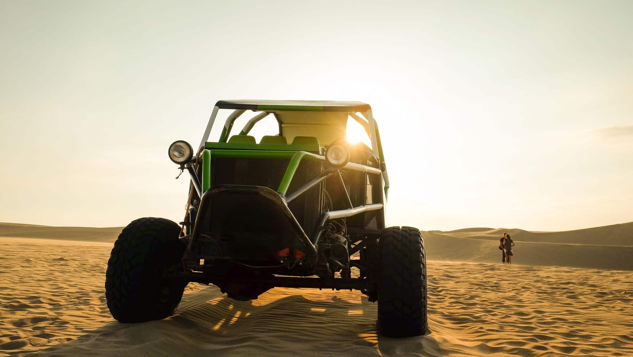 Dune buggy in the desert surrounded by sand at sunset hour. Young couple in the background. Selective focus, space for copy. Huacachina, Ica, Peru. Adventure, travel, journey concept landscape.