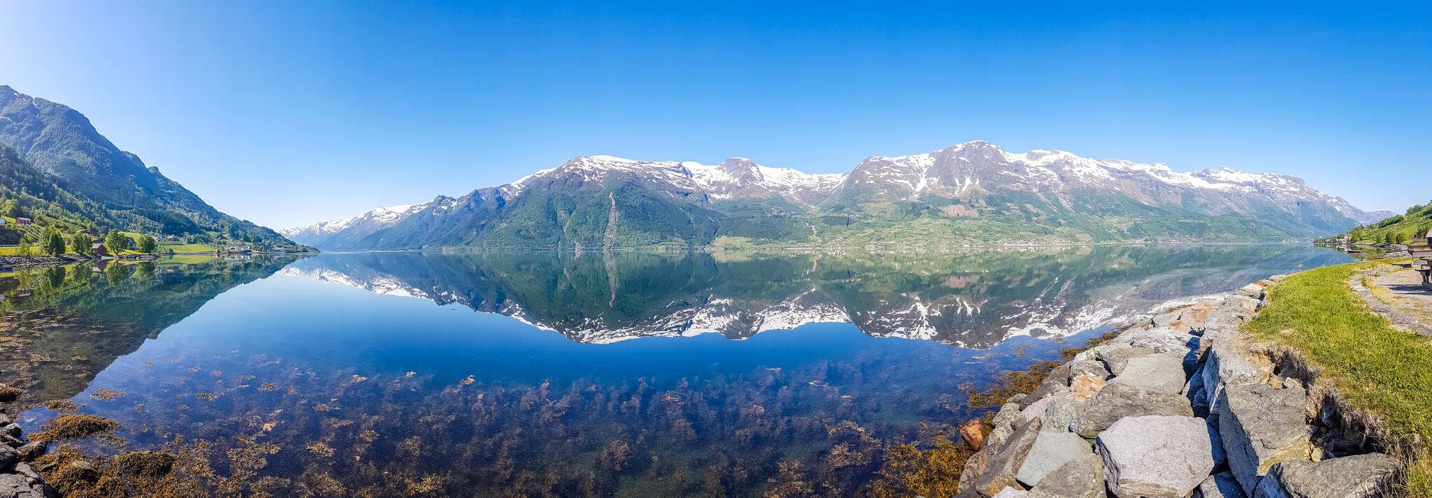An endless chain of mountains reflecting itself in a calm water of Eidfjord. Taller parts of the mountains are partially covered with snow. Sunny and bight weather, clear blue sky. Romantic landscape