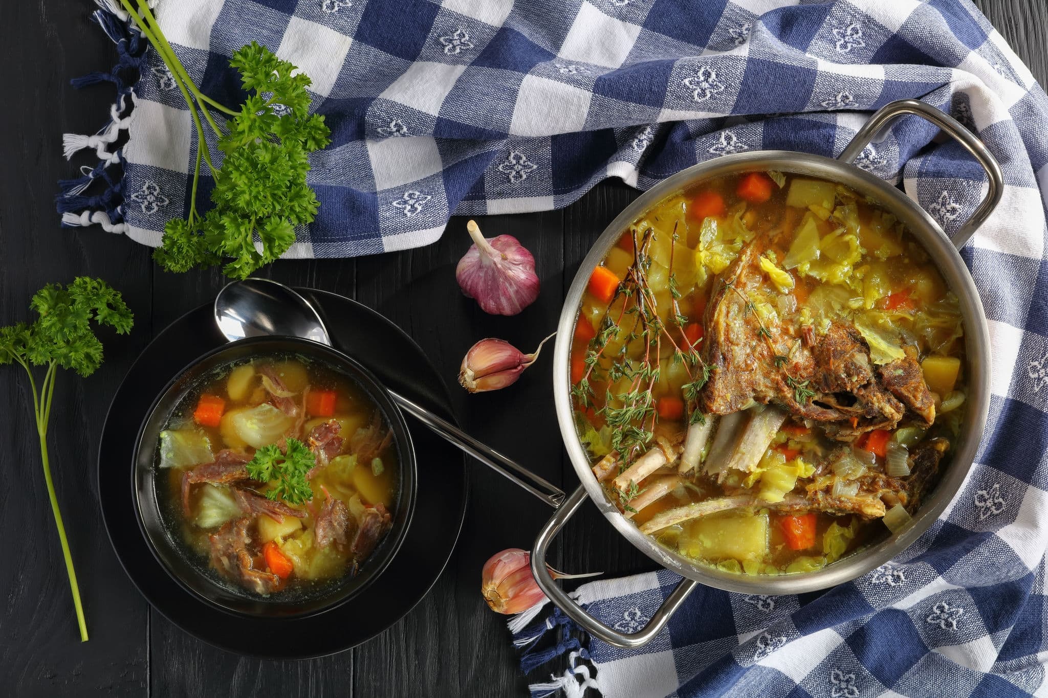 close-up of delicious Icelandic Lamb Soup with vegetables and spices or kjotsupa in a stainless steel casserole pan  and in bowl on wooden table with kitchen towel, traditional recipe, view from above