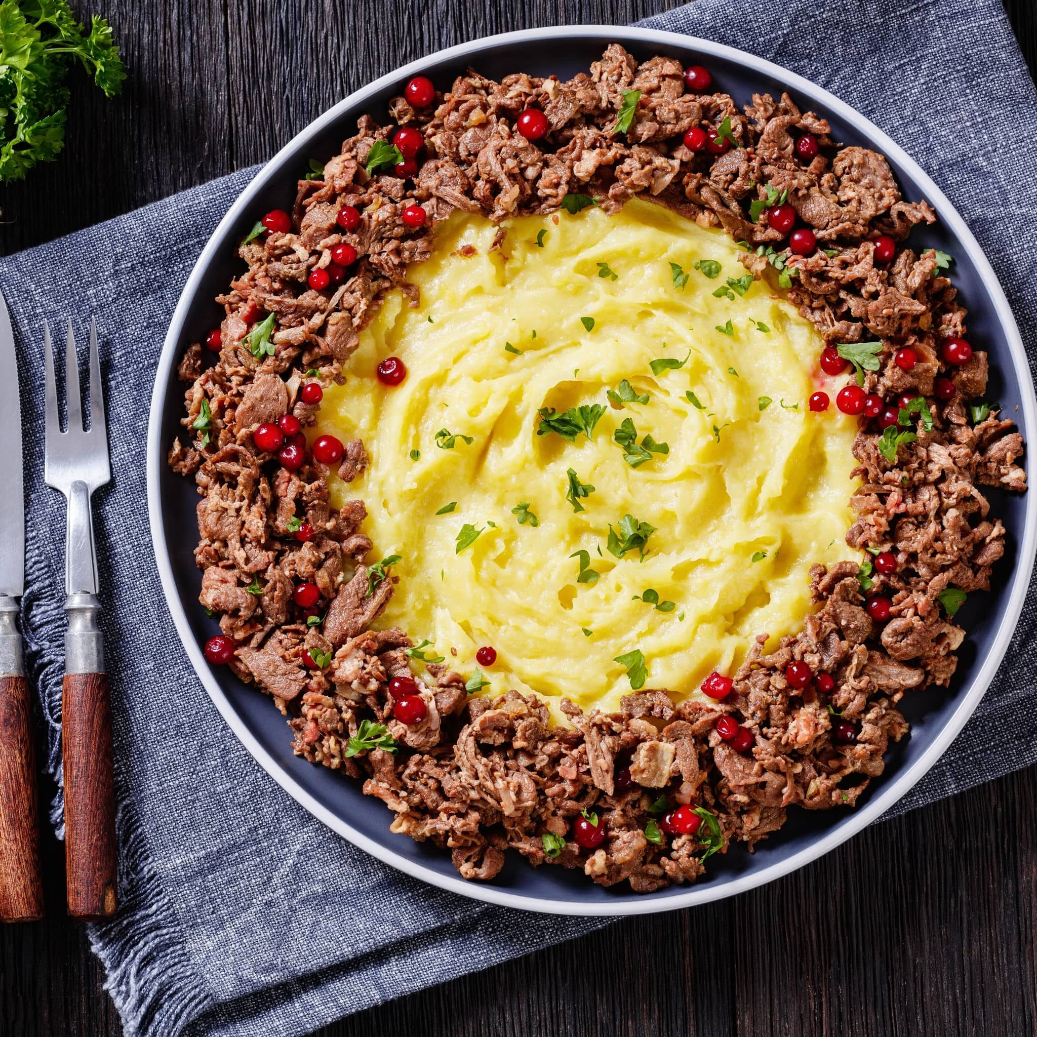 Reindeer Stew with Lingonberries and potato puree on plate on wood table with ingredients and pickles on background, finnish cuisine,  horizontal view from above, flat lay, close-up