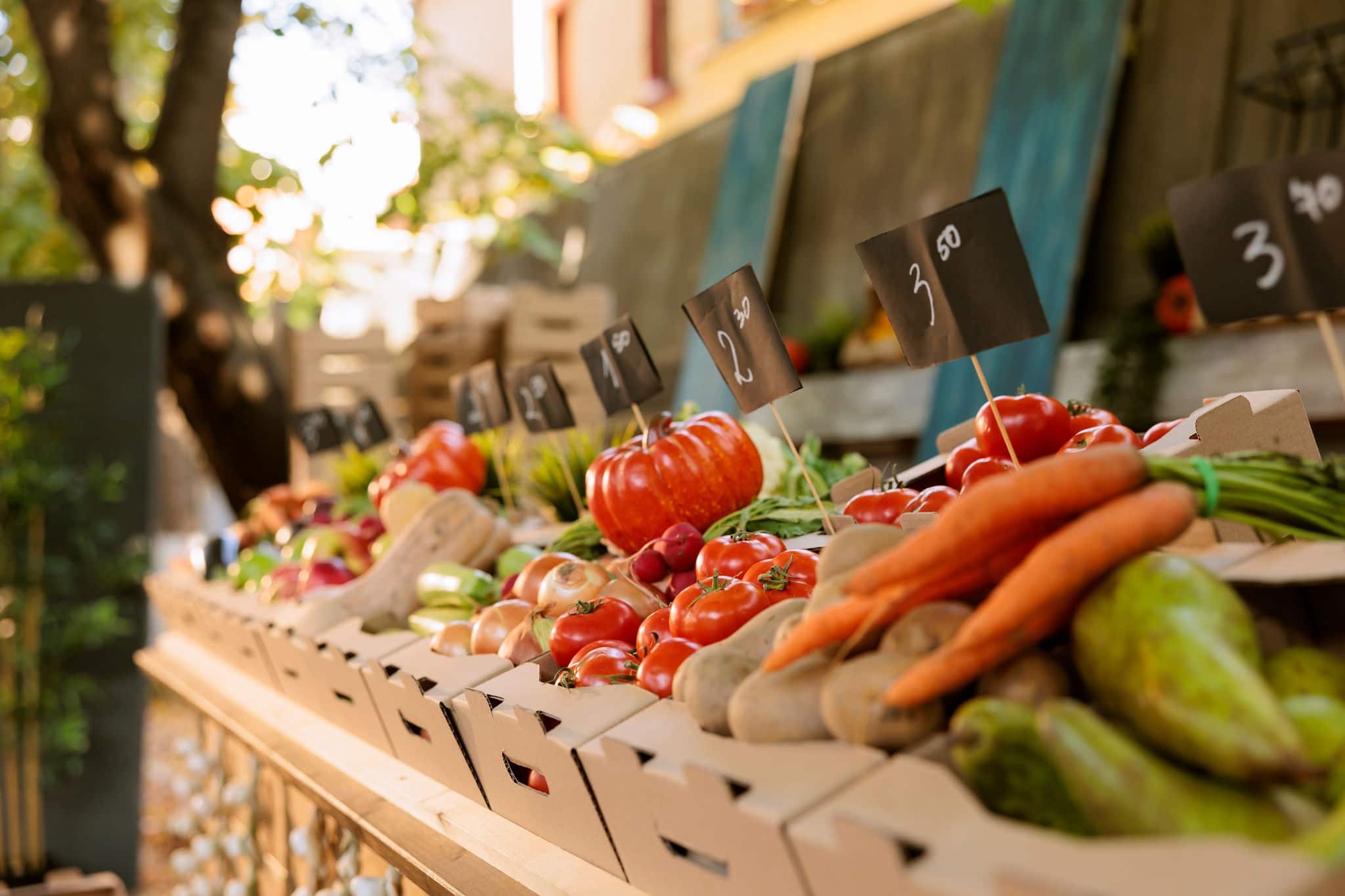 Detailed view of organic agricultural fresh produce in cardboard boxes placed on greenmarket stands with price tags. Closeup of locally grown colorful fruits and vegetables at farmers market booth.