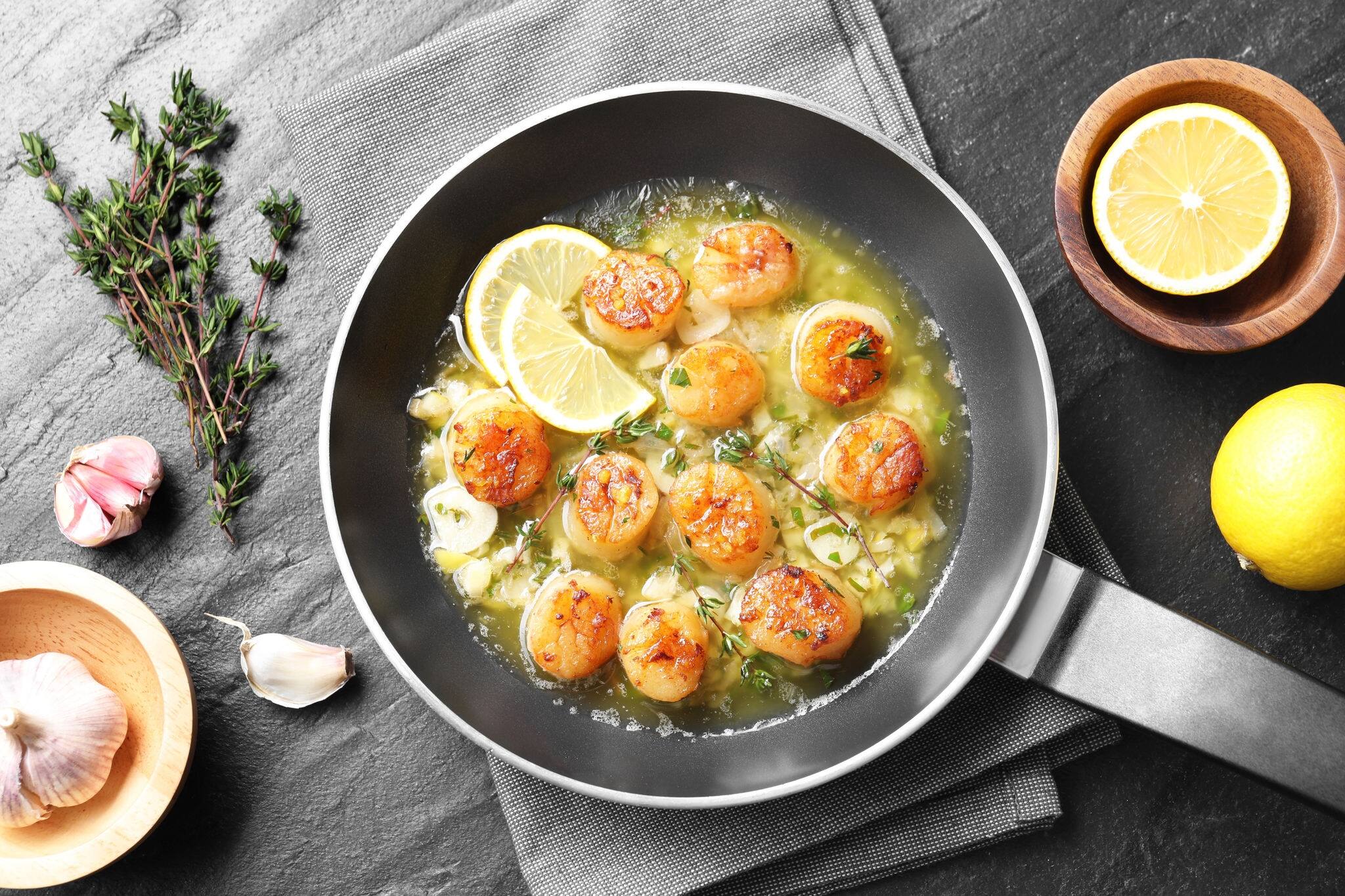 Delicious scallops with sauce in frying pan and ingredients on dark gray textured table, flat lay