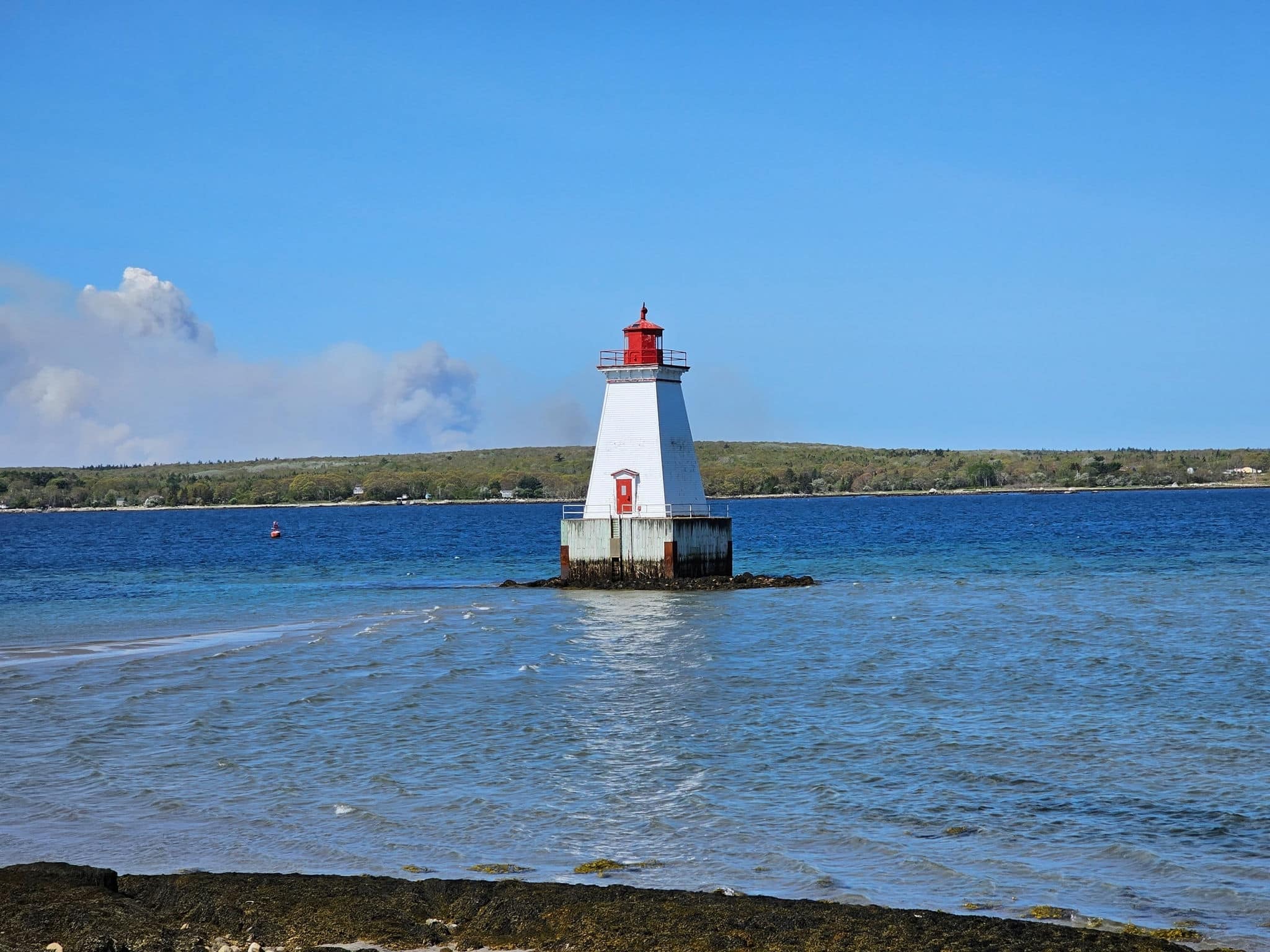 A lighthouse sitting in the middle of the water with a smoke cloud from a wildfire off to the side.