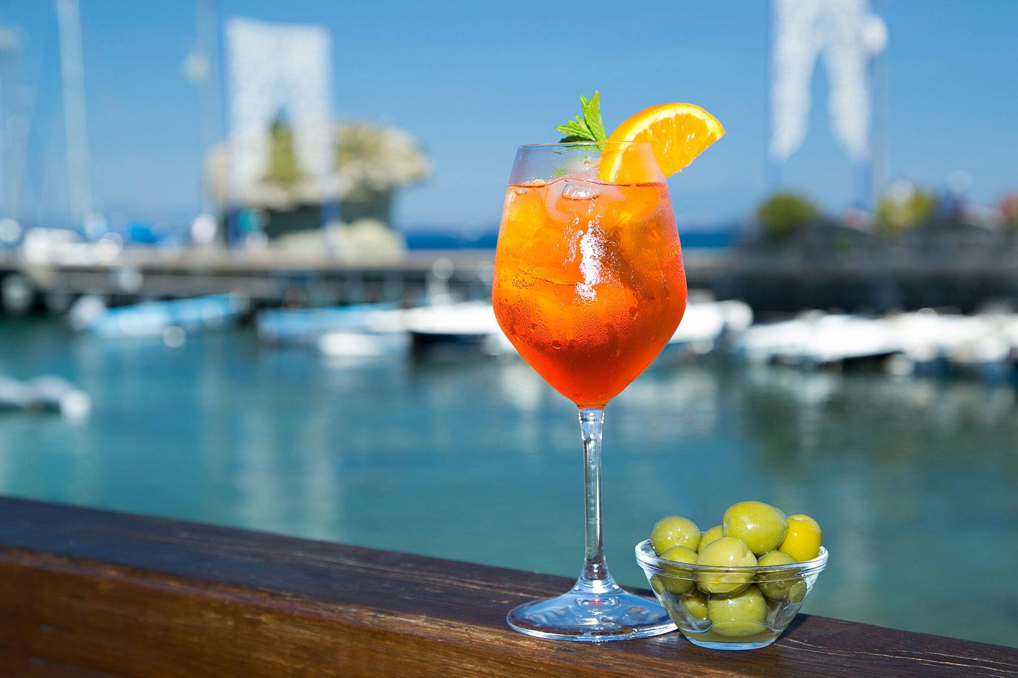 Cold glass of aperol spritz wit olive appetizer stand near the sea and boats