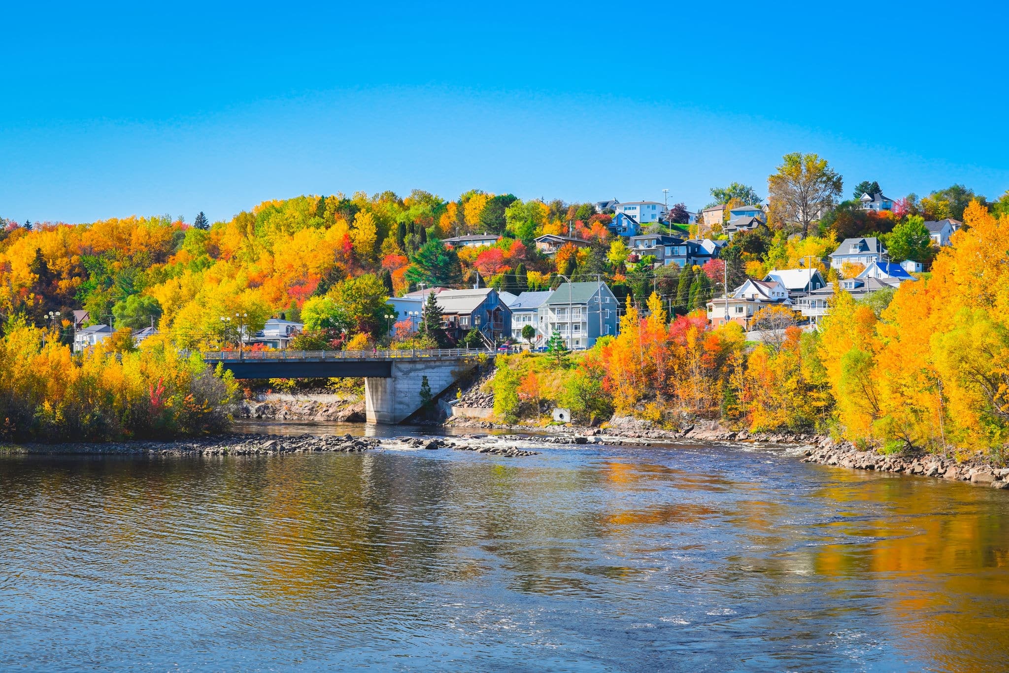 Beautiful autumn landscape view in Chicoutimi, Saguenay, Quebec City, Canada. A village on shore of lake with colorful trees, river and bridge against blue sky reflected in water during fall season.