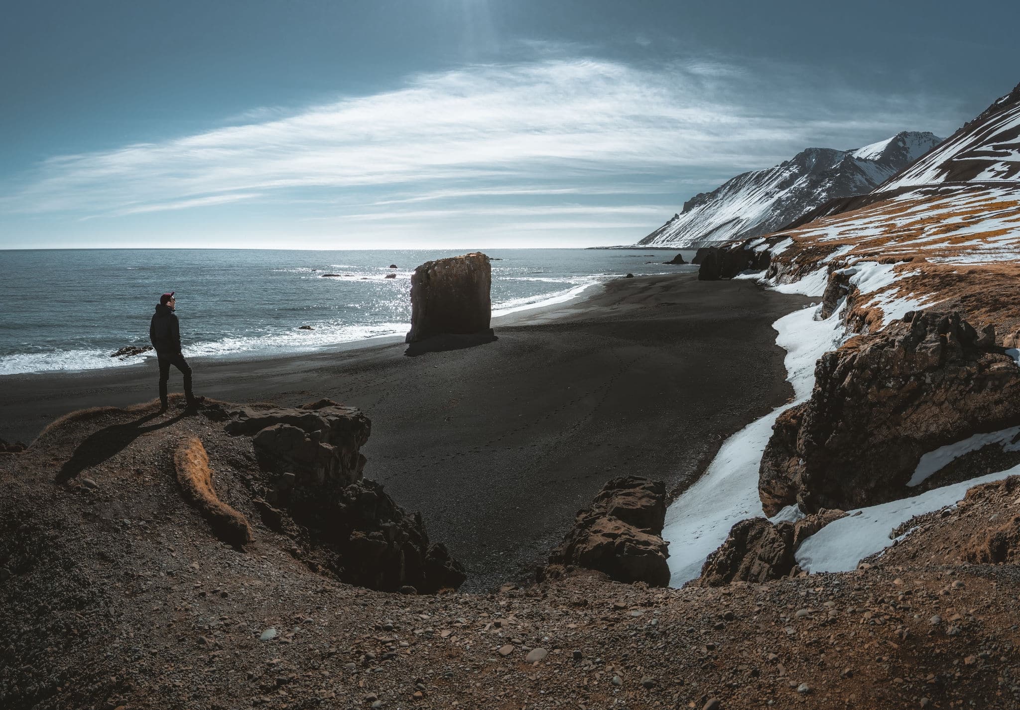 Person standing at black sand beach in Iceland. Fauskasandur near village of Djupivogur. Black sand beachwith snow and beautiful sunny weather. Seastack monolith.