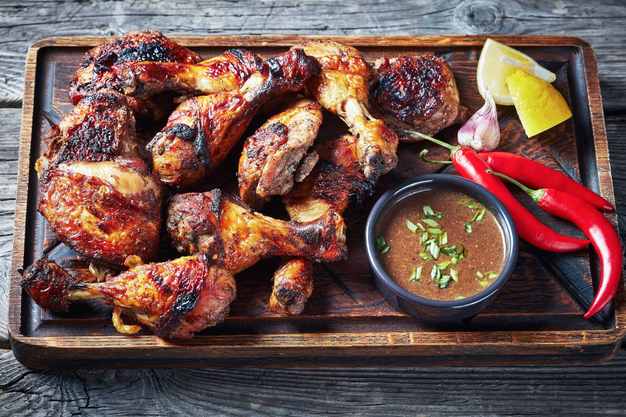 hot Grilled Jamaican Jerk Chicken on a rude board with sauce and lemon on a rustic wooden table, horizontal view from above, close-up
