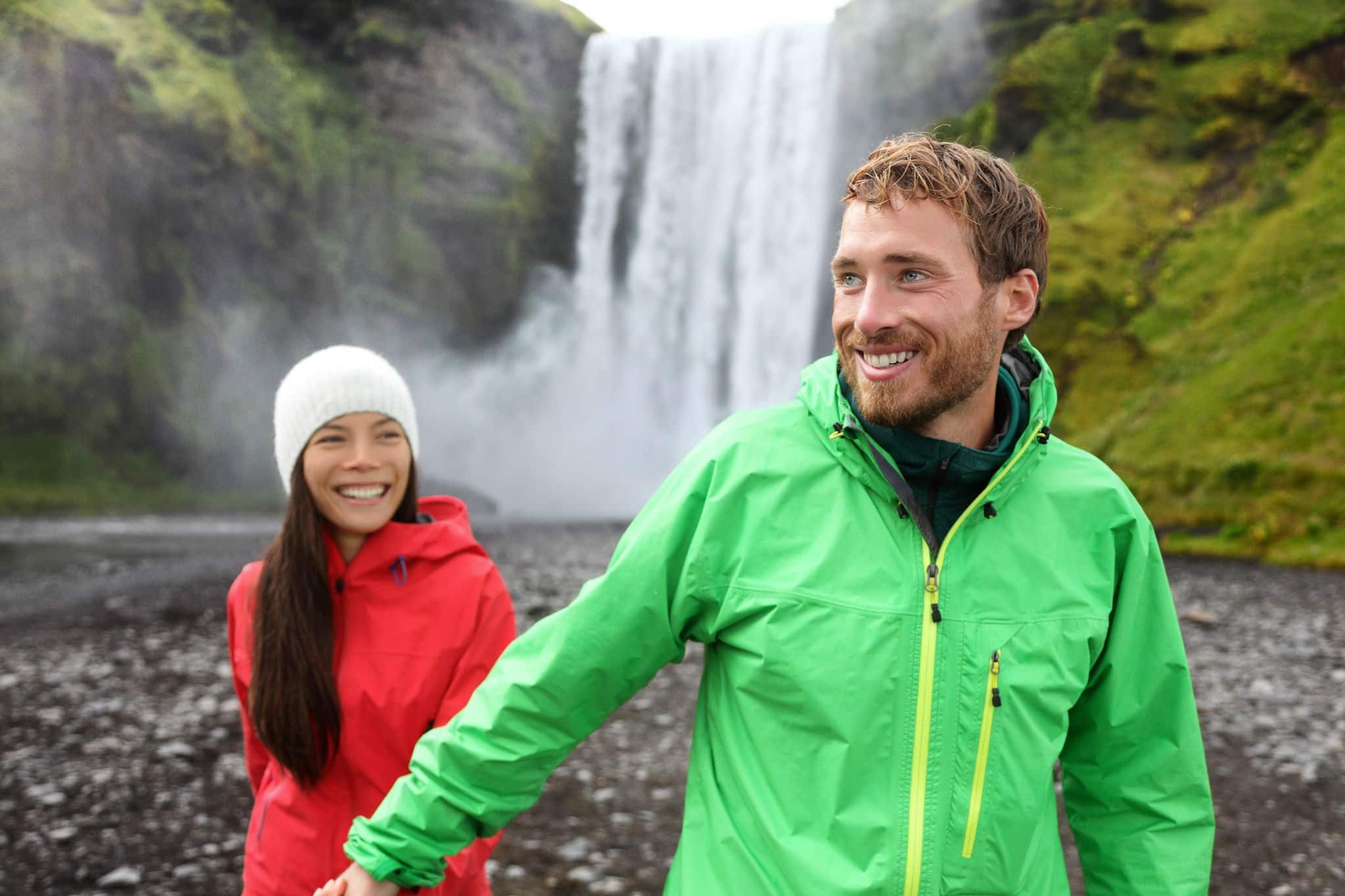 Happy couple holding hands by waterfall outdoors in front of Skogafoss on Iceland. Couple visiting famous tourist attractions and landmarks in Icelandic nature landscape on Golden Circle.