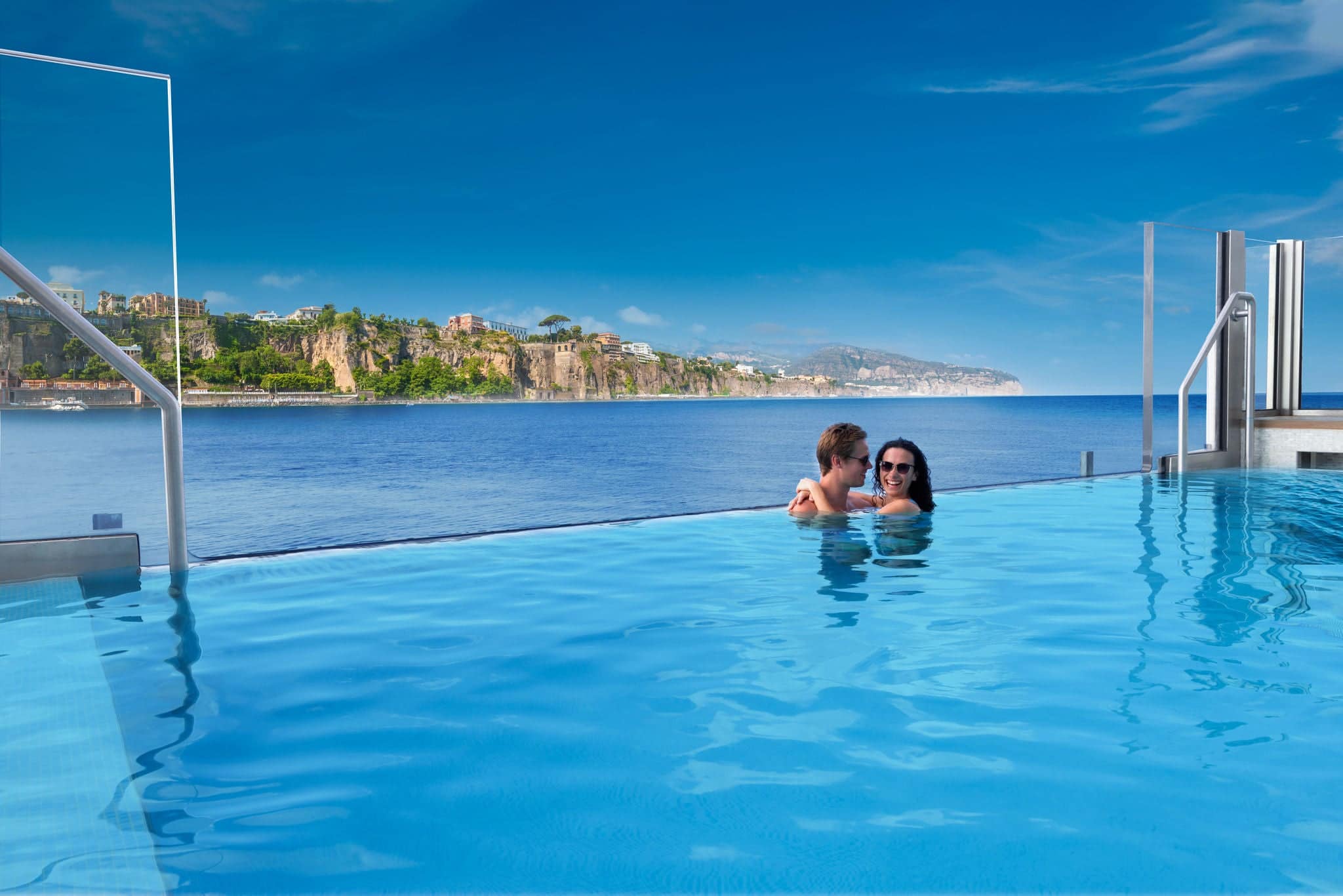 a couple in an infinity pool with open water and a coastline in the background
