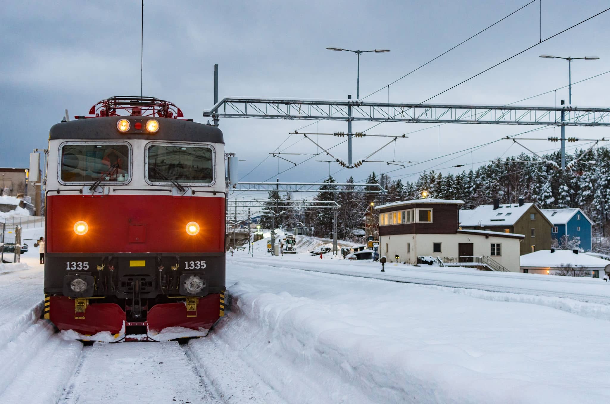 Red train in a snowy and cold train station next to the platform, station and trees in the background.