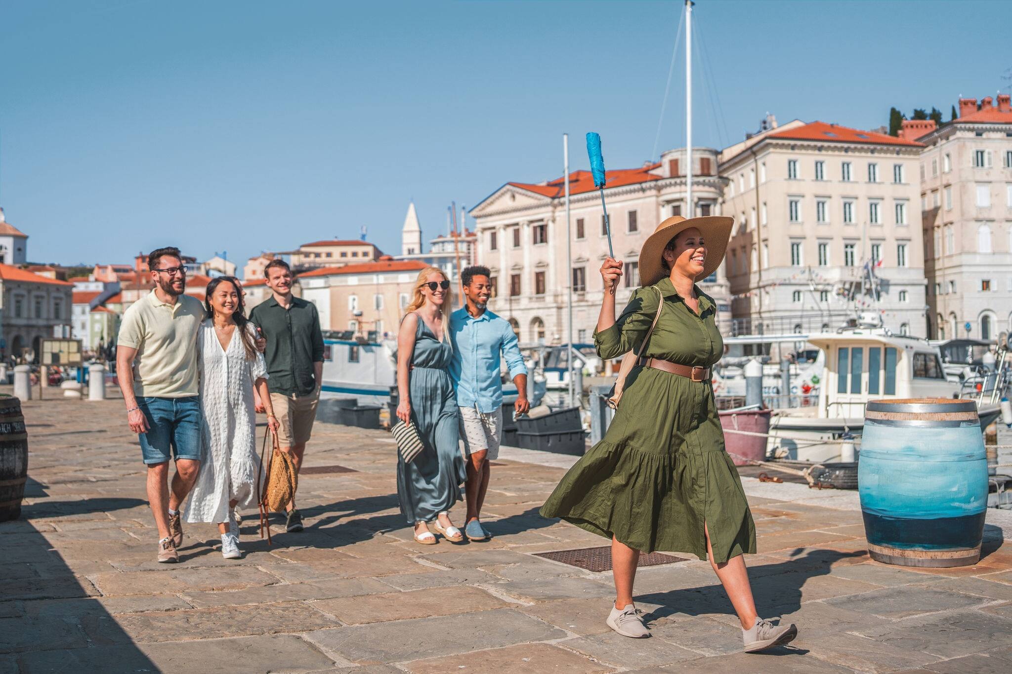 Diverse group of travelers, led by a mixed-race female tour guide, as they explore a small Mediterranean town. They are engaged in the exploration, discovering the local charm and having a great time.