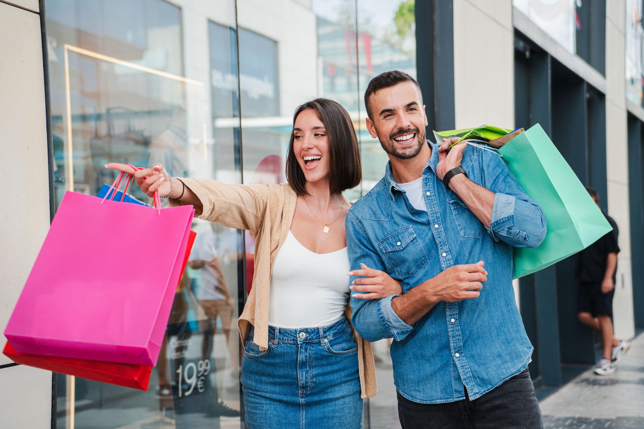 A couple points at the display of a store in the mall, deciding whether to buy an offer. A customer man and a client woman with shopping bags, smiling and looking at the shop window to purchase a gift