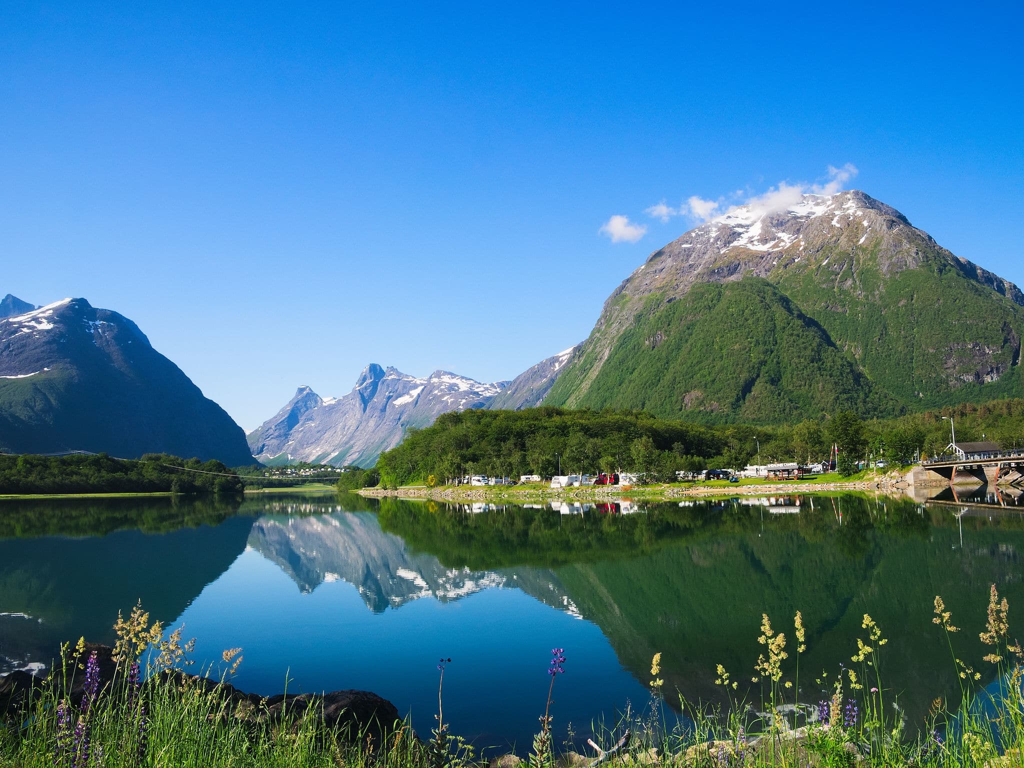 Fabulous Norway Landscape includes beautiful mountains with snow, forest and bare rock, a bridge, an RV park and blue sky reflected in the Rauma River near Andalsnes, Norway.