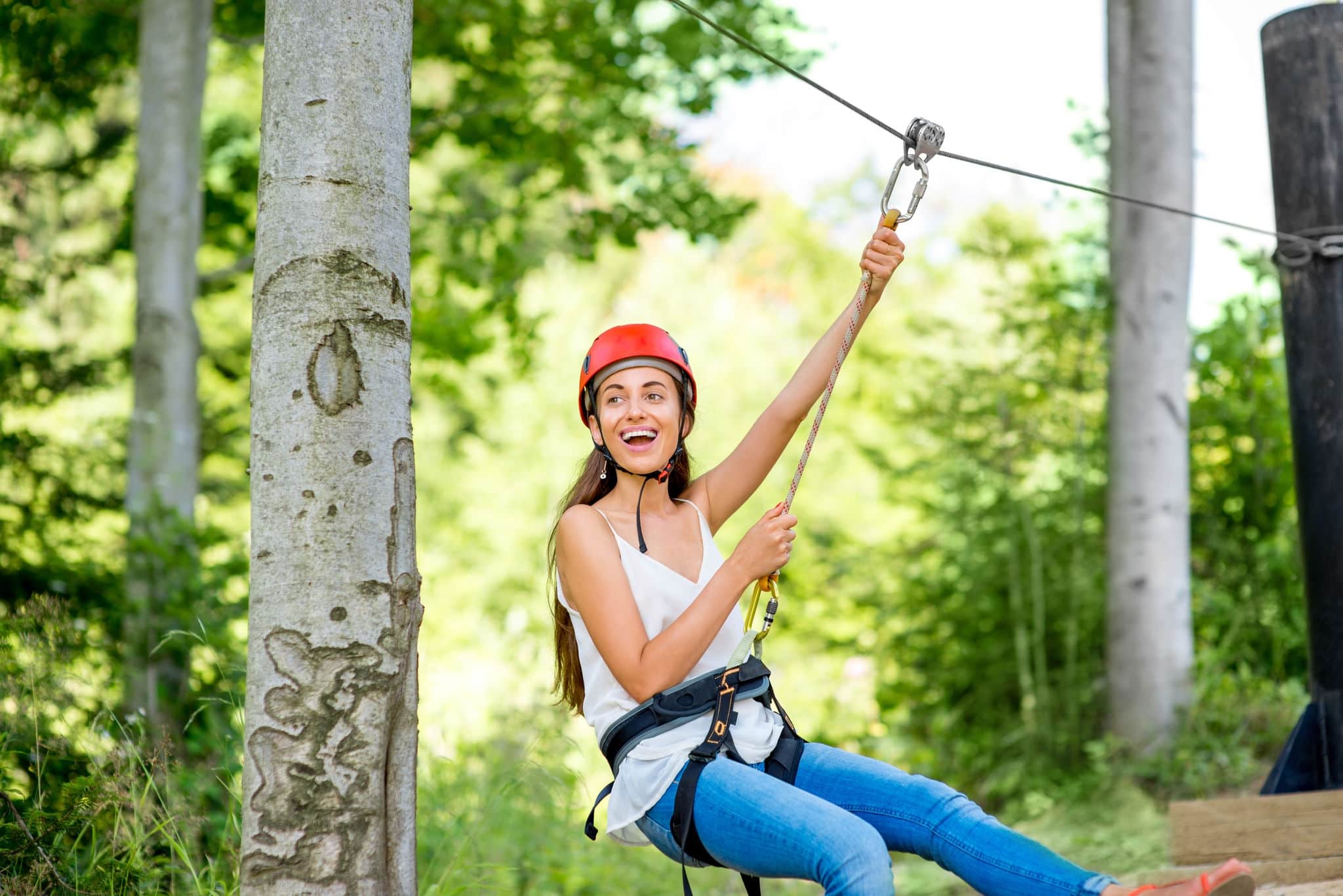 Young and pretty woman in red helmet riding on a zip line in the forest. Active sports kind of recreation