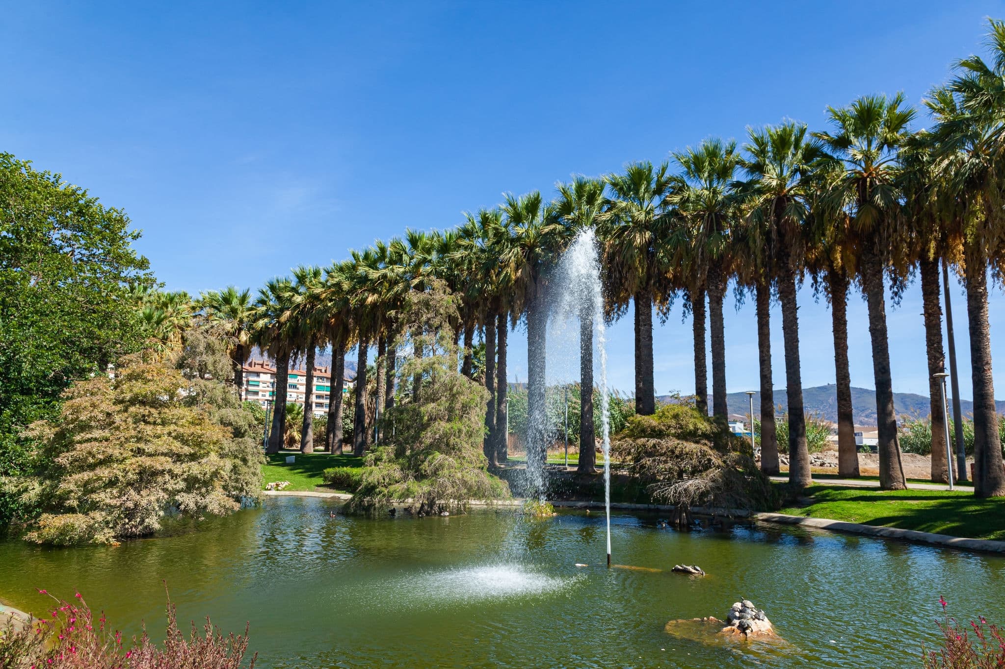 Pueblos de America Park in Motril, Spain, with a fountain spouting water in the middle of a lake and tall palm trees.