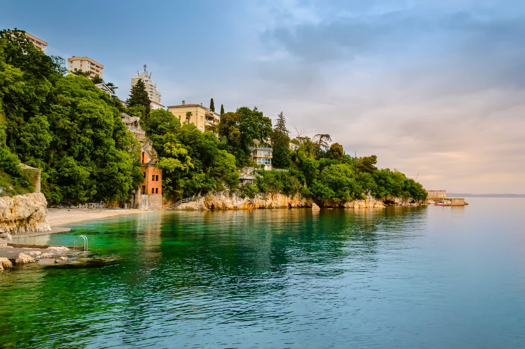 Glavanovo beach in Rijeka, Croatia at sunset. Stunning croatian coast with small beach, buildings, ruins and rocky cliffs covered with lush green trees. Steep stairway leads from hill down to sea.