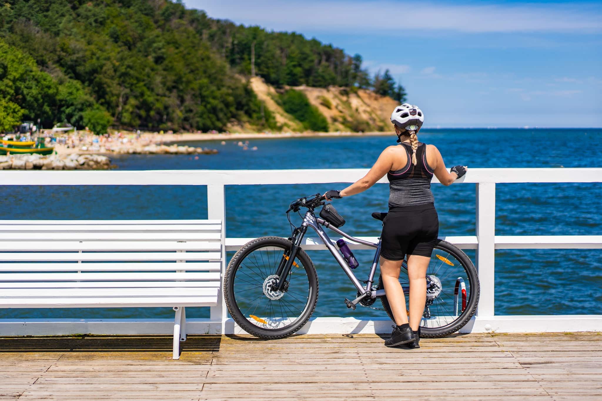 Pier in Orlowo Gdynia Poland. View from pier to Orlowski Cliff. Bike trip around Tri-City. Woman dressed in sportswear and white bike helmet standing backwards by bike on pier and looking at cliff.