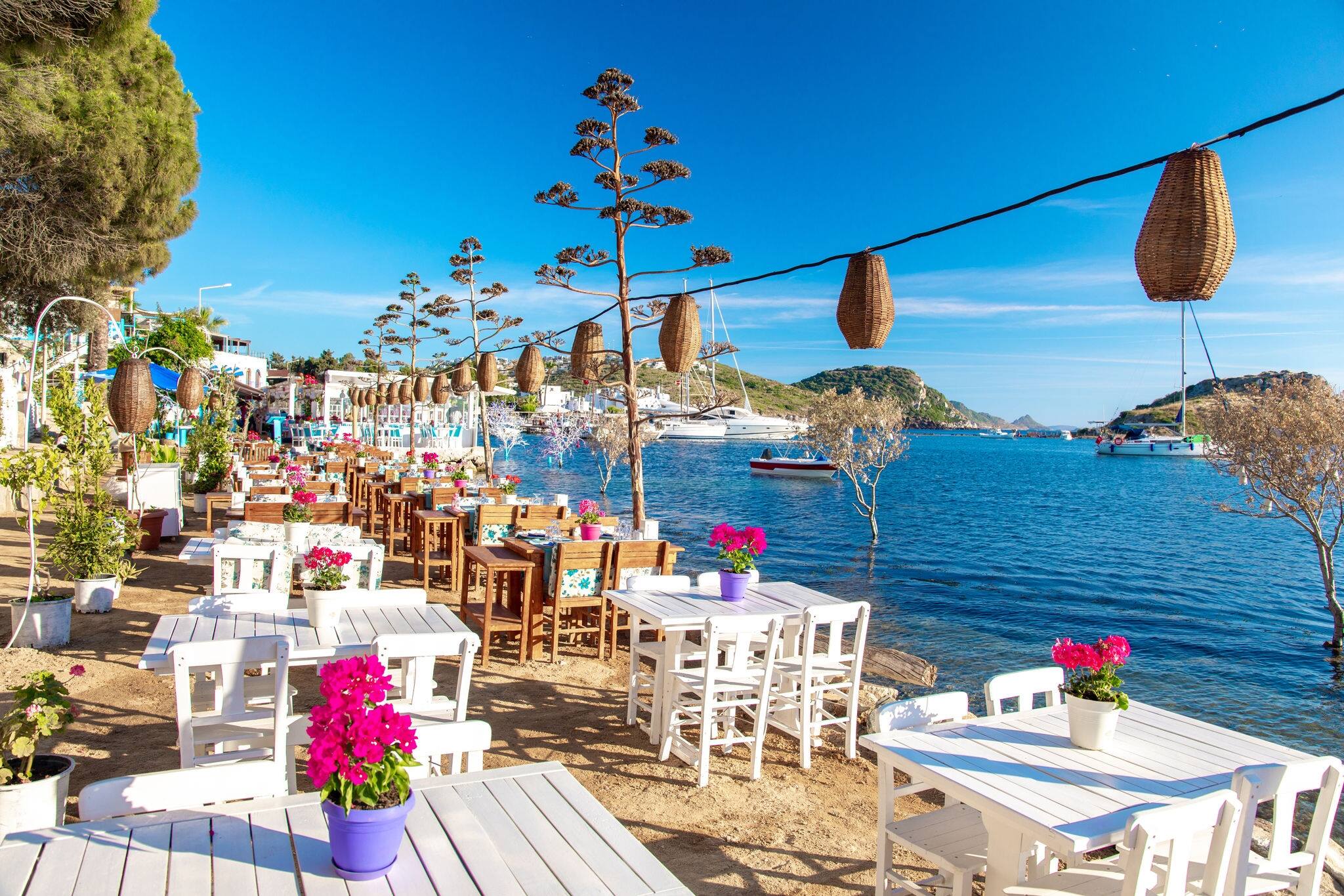 View of restaurant or cafe and bougainvillea flowers on beach in Gumusluk, Bodrum city of Turkey. Aegean seaside style colorful chairs, tables and flowers in Bodrum town near beautiful Aegean Sea.