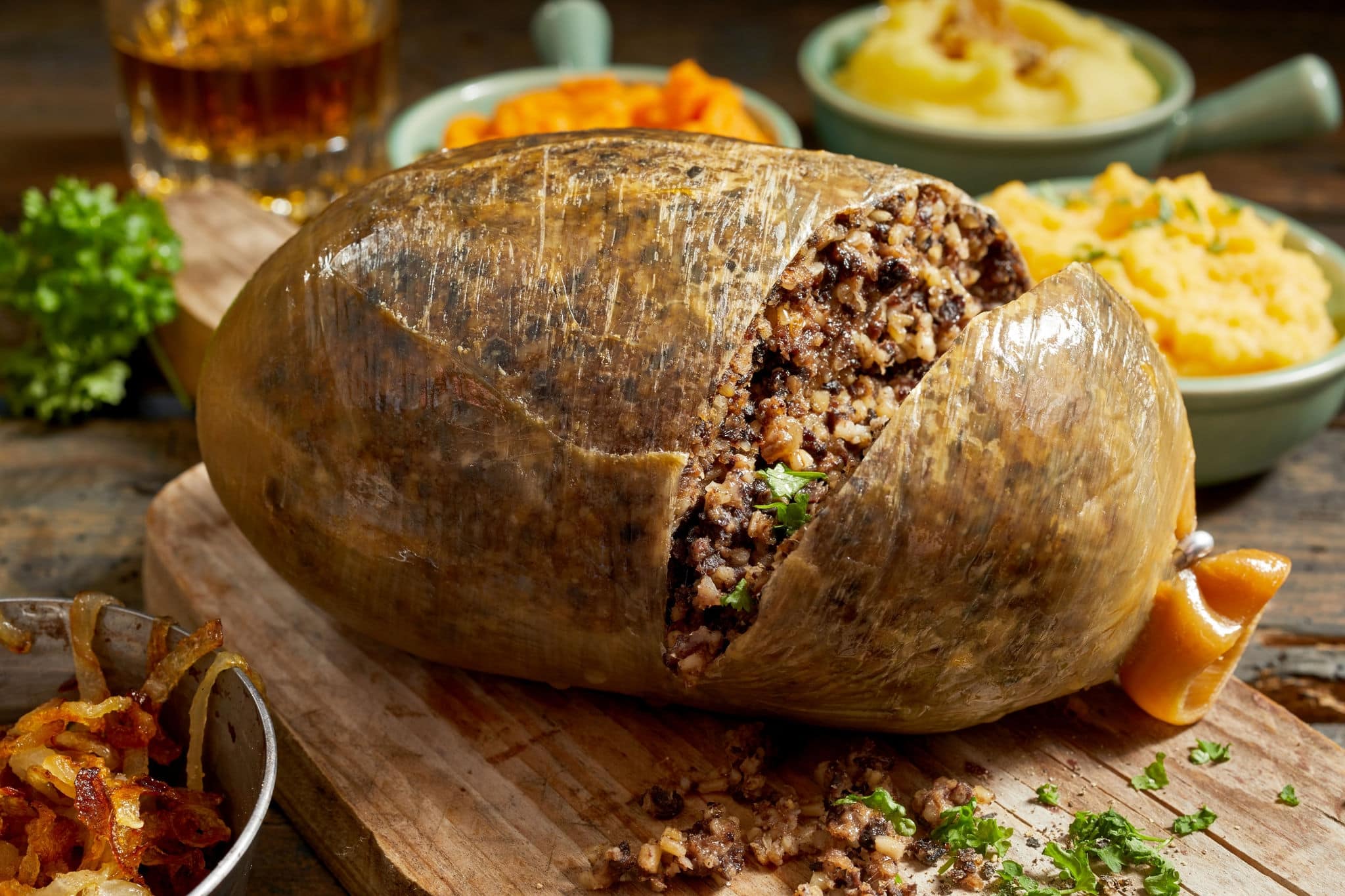 Sliced open cooked Scottish haggis showing the minced texture of the meat mixture on a wooden board with side dishes of mashed potato, turnip and carrot with fresh herbs