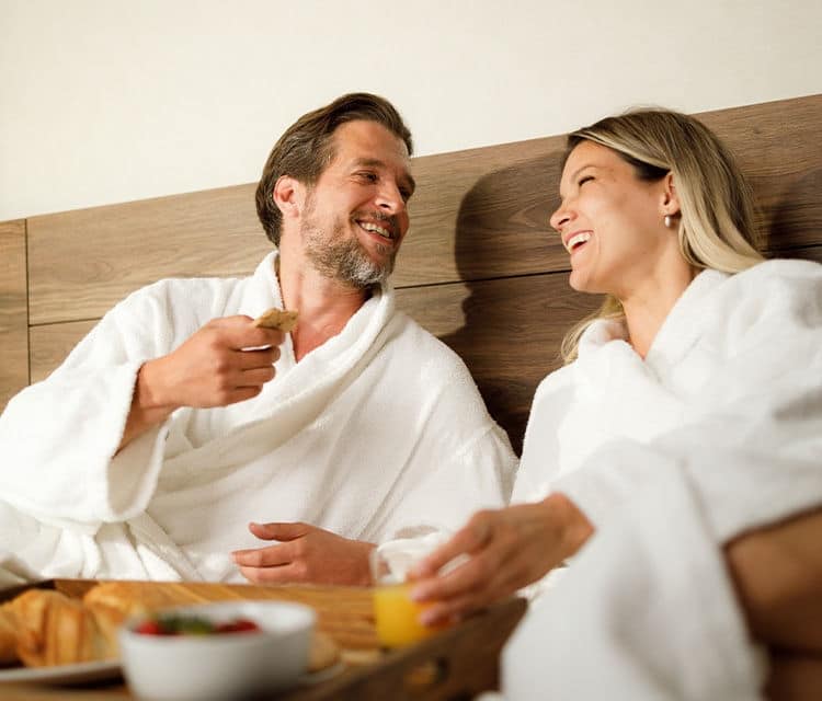 A couple in white robes relaxes with a breakfast tray.