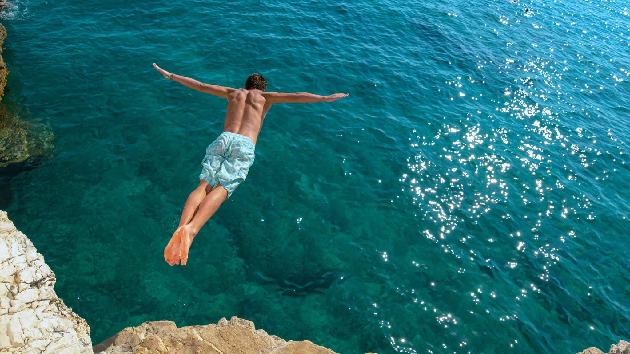 COPY SPACE: Athletic young man jumping off a rocky ledge and into the glistening blue ocean. Unrecognizable male tourist on a relaxing summer vacation in does cliff diving on a beautiful sunny day.
