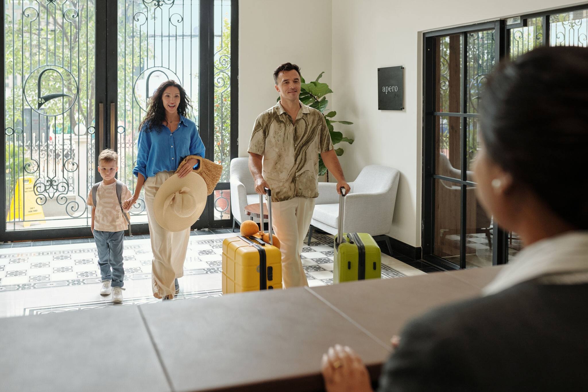 Young family entering hotel lobby carrying luggage, being greeted by receptionist at front desk. Smiling parents accompanied by small child holding hands